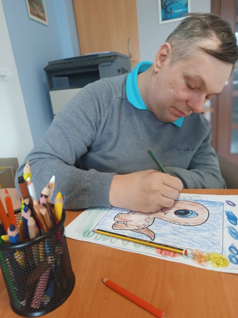 Podopieczny koloruje kredkami obrazek z dinozaurem