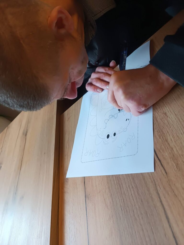 Podopieczny koloruje kredkami obrazek z dinozaurem